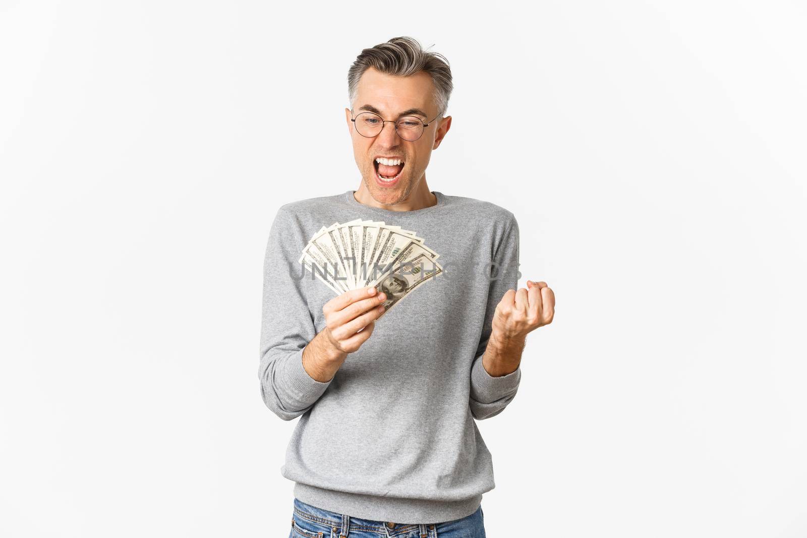 Image of cheerful middle-aged man in glasses and gray sweater, winning money, holding cash and rejoicing, standing delighted over white background.