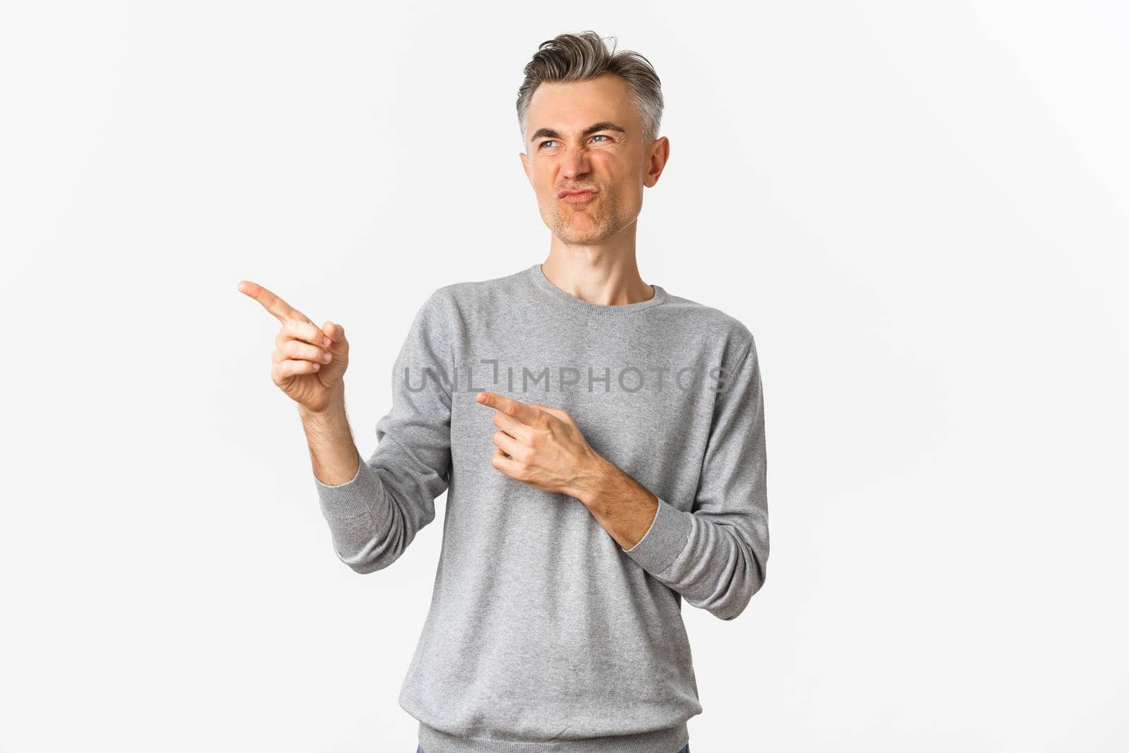 Portrait of skeptical handsome adult man, wearing grey casual sweater, grimacing as looking and pointing at something bad, express negative opinion, standing over white background.