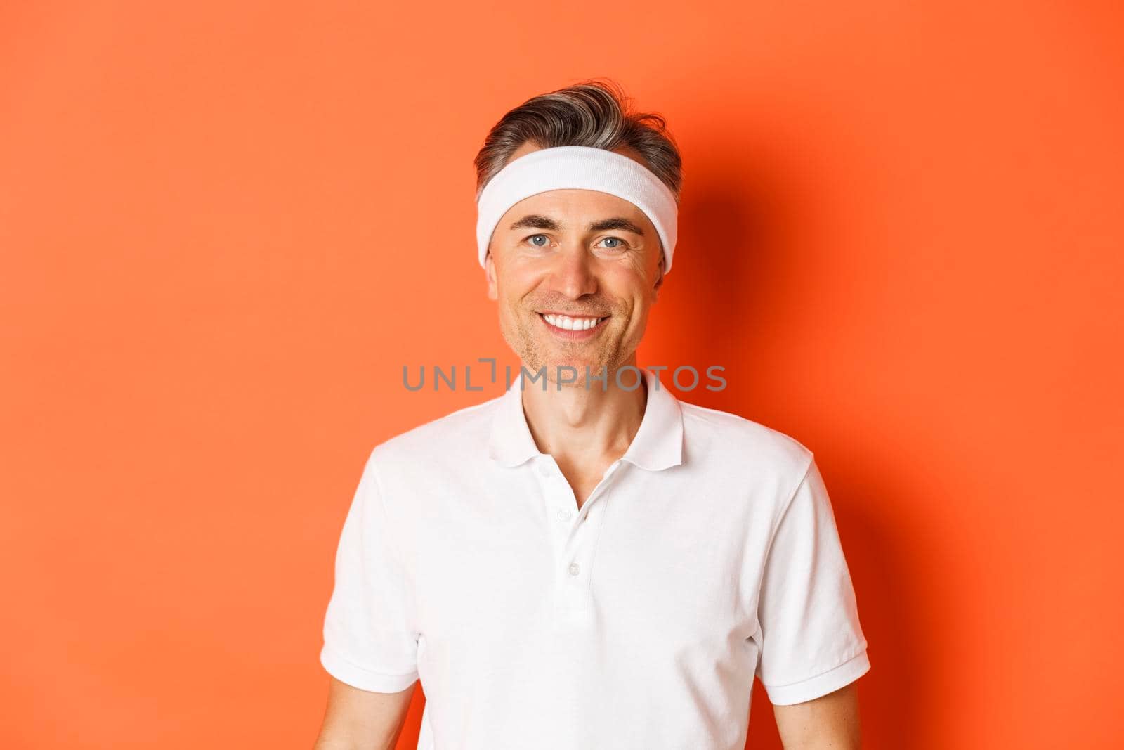 Close-up of handsome and fit middle-aged sportsman, wearing headband and white t-shirt, smiling happy, standing over orange background.