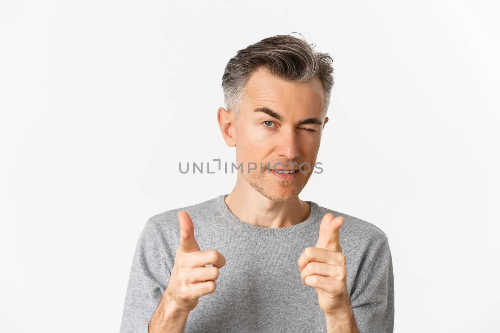 Close-up of handsome cheeky man with gray hair, winking and pointing fingers at camera, praising you, well done gesture, standing over white background.