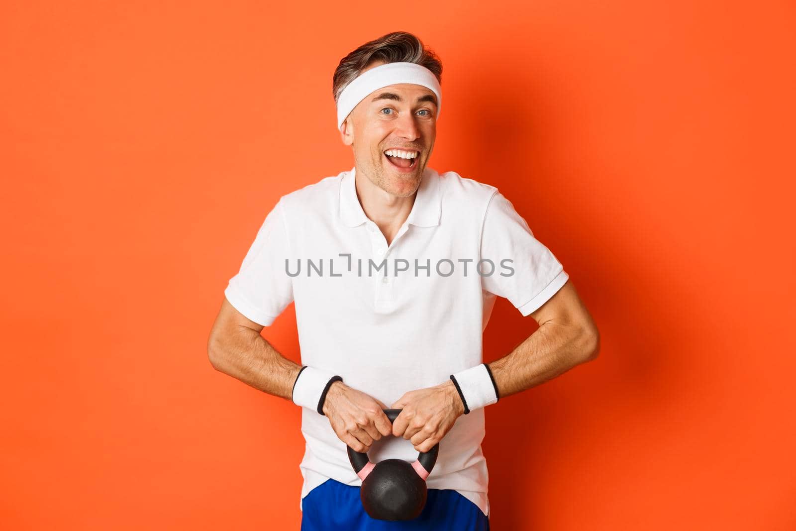 Concept of workout, gym and lifestyle. Image of handsome middle-aged guy doing sport exercises, lifting kettlebell and smiling, standing over orange background.