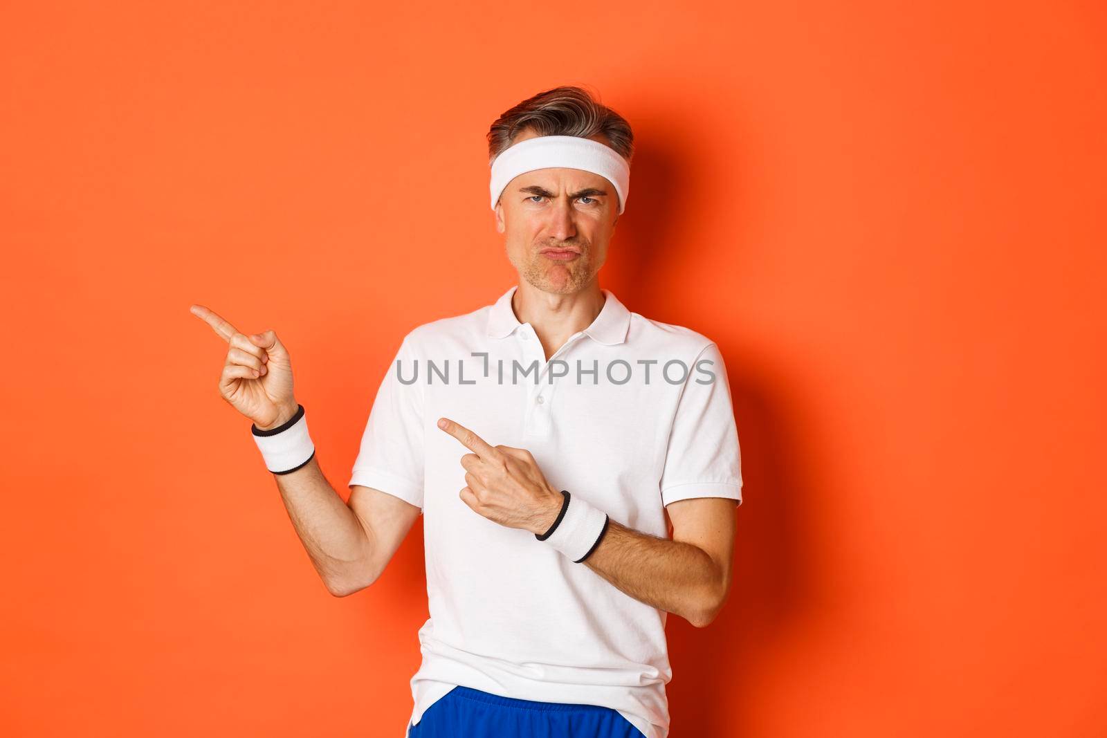 Portrait of disappointed middle-aged man in sportswear, complaining and pointing fingers left at logo, showing something bad, standing over orange background.