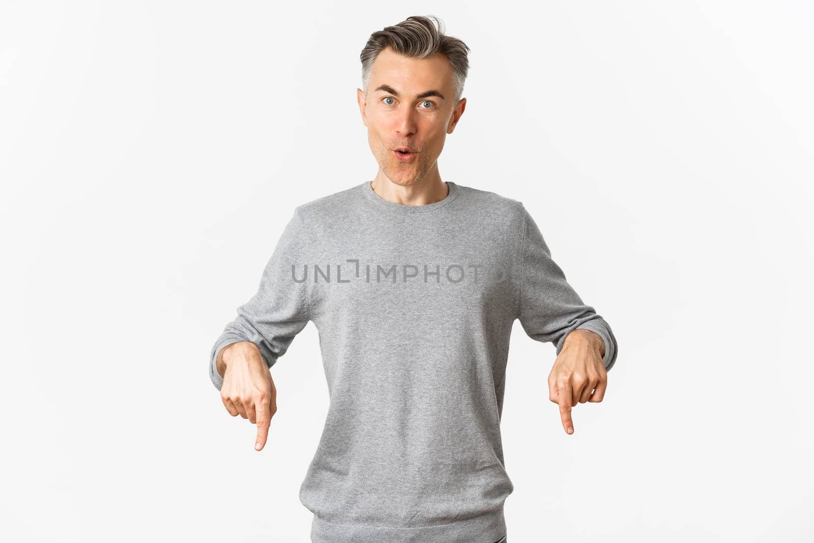Surprised handsome middle-aged guy in grey sweater asking question about product, pointing fingers down and looking amazed, standing over white background.