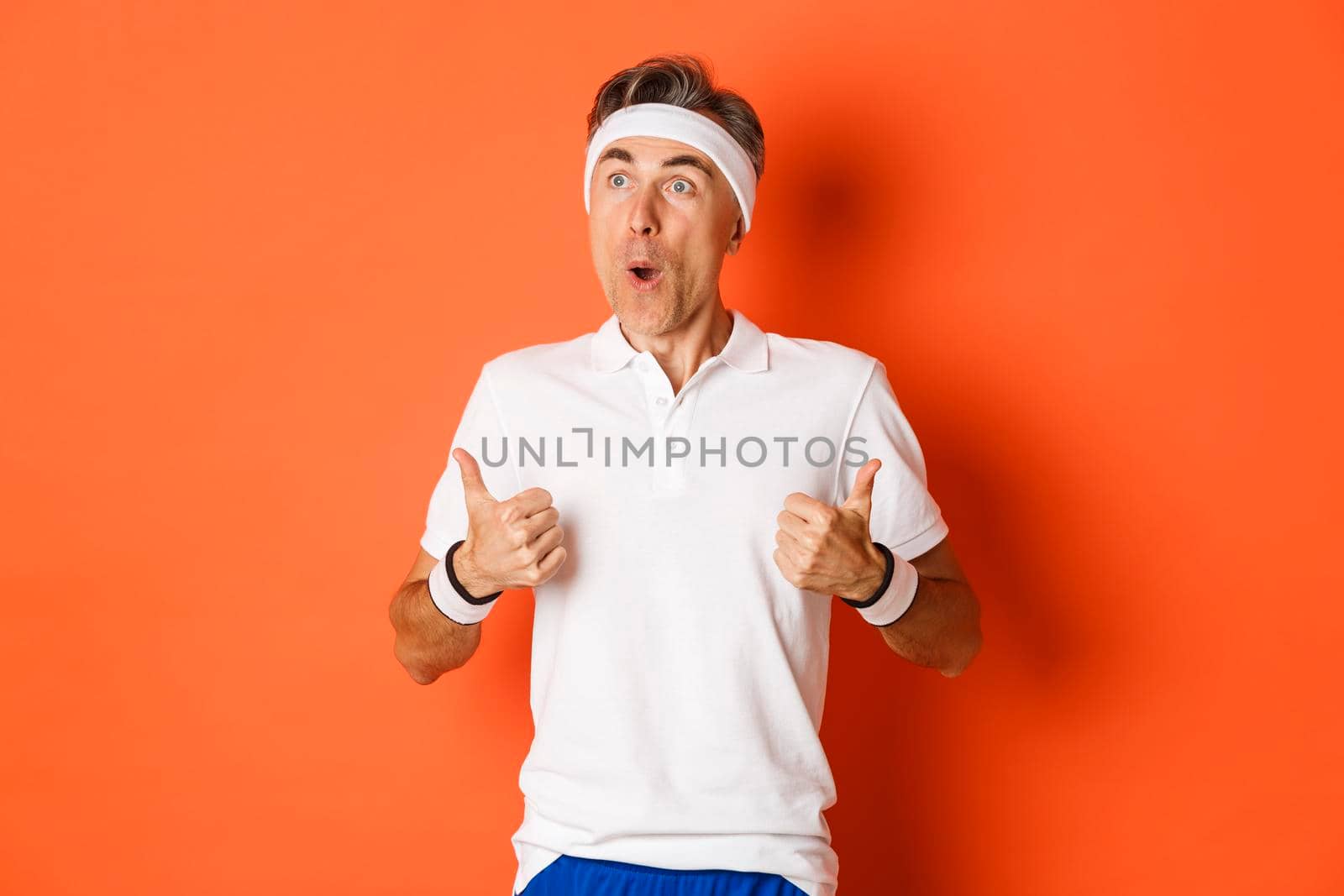 Concept of sport, fitness and lifestyle. Portrait of amazed and excited middle-aged sportsman, looking at upper left corner at something awesome, showing thumbs-up, orange background.