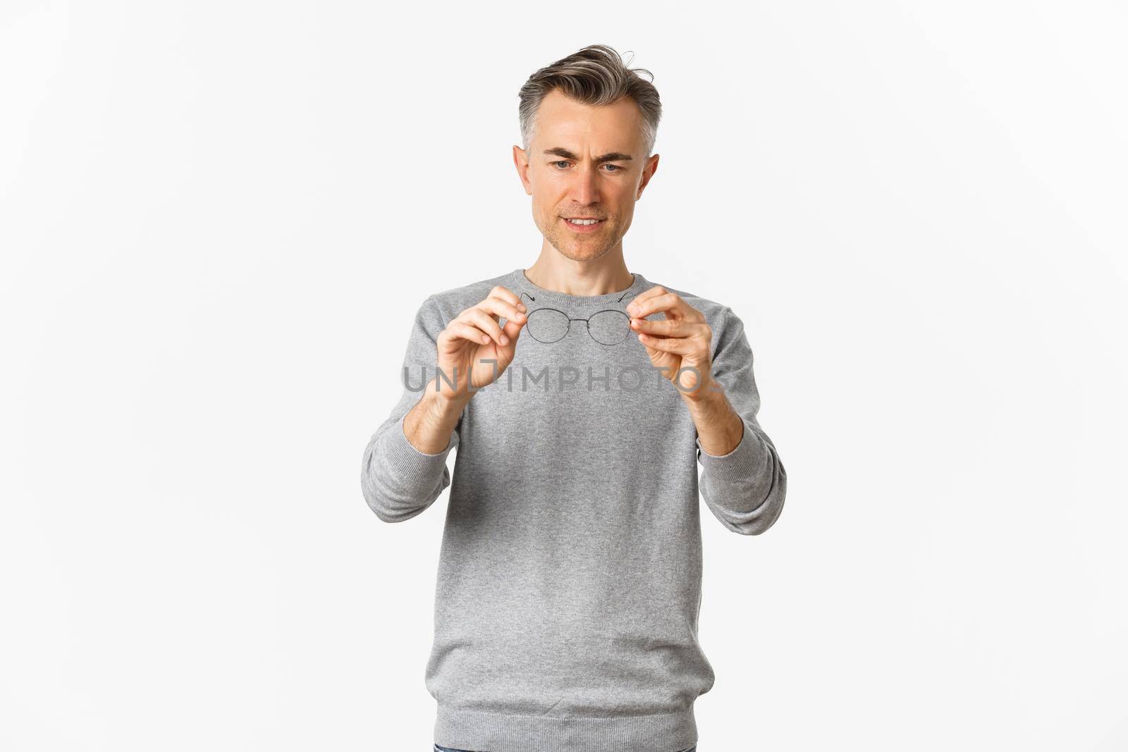 Portrait of middle-aged man in grey sweater, looking at glasses, standing over white background.