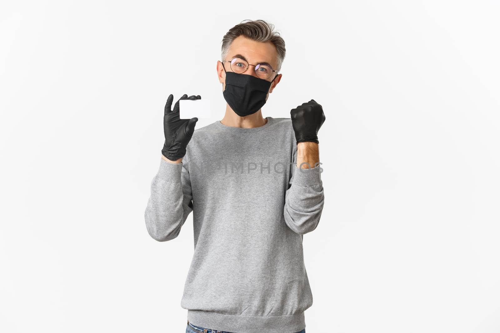 Concept of covid-19, social distancing and lifestyle. Image of attractive middle-aged man in medical mask, gloves and glasses, showing credit card and rejoicing, standing over white background.