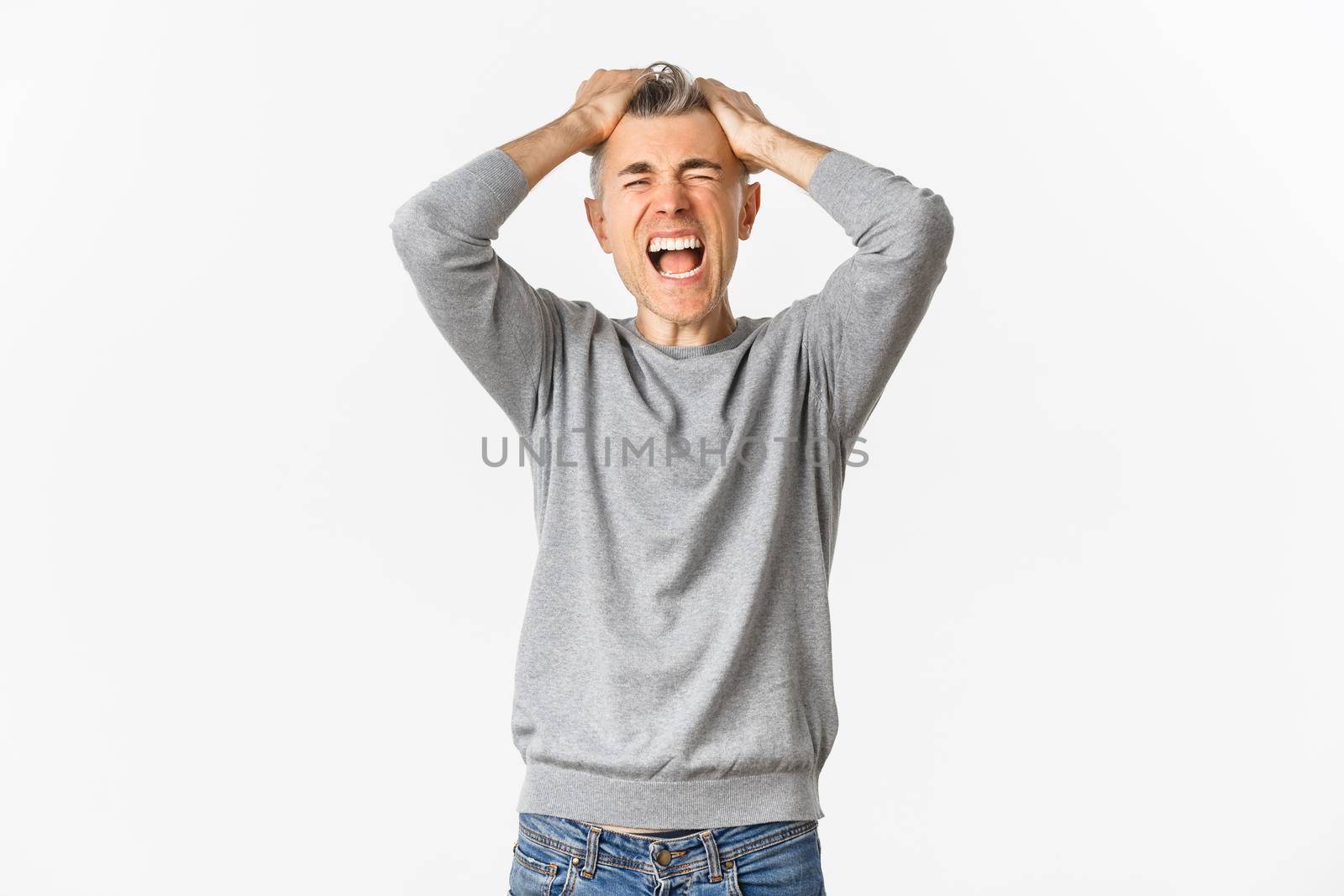 Image of frustrated middle-aged man panicking, losing something, ripping hair on head and grimacing from distress and failure, standing over white background.