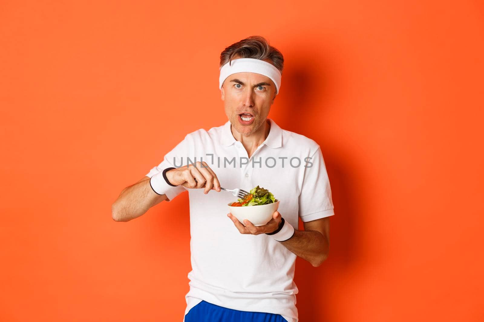 Portrait of angry middle-aged male athlete, eating salad and looking disturbed, standing over orange background.