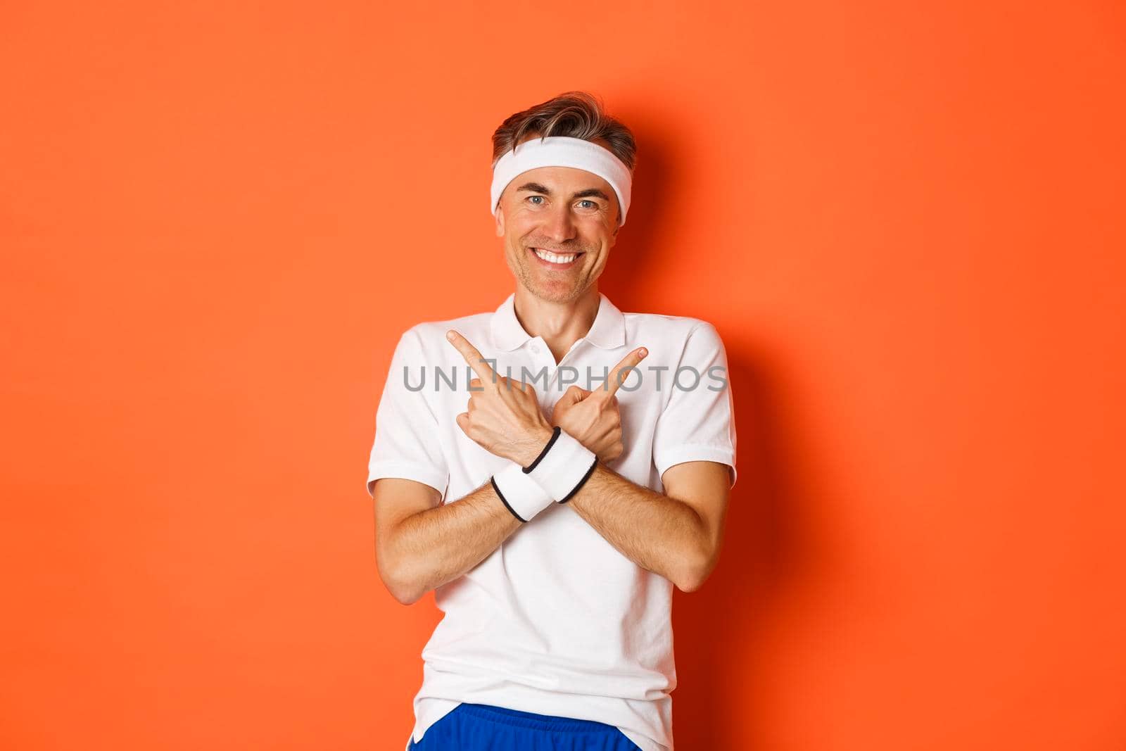 Concept of sport, fitness and lifestyle. Portrait of happy, handsome middle-aged guy in workout clothes, pointing fingers sideways, showing left and right promo banners, orange background.