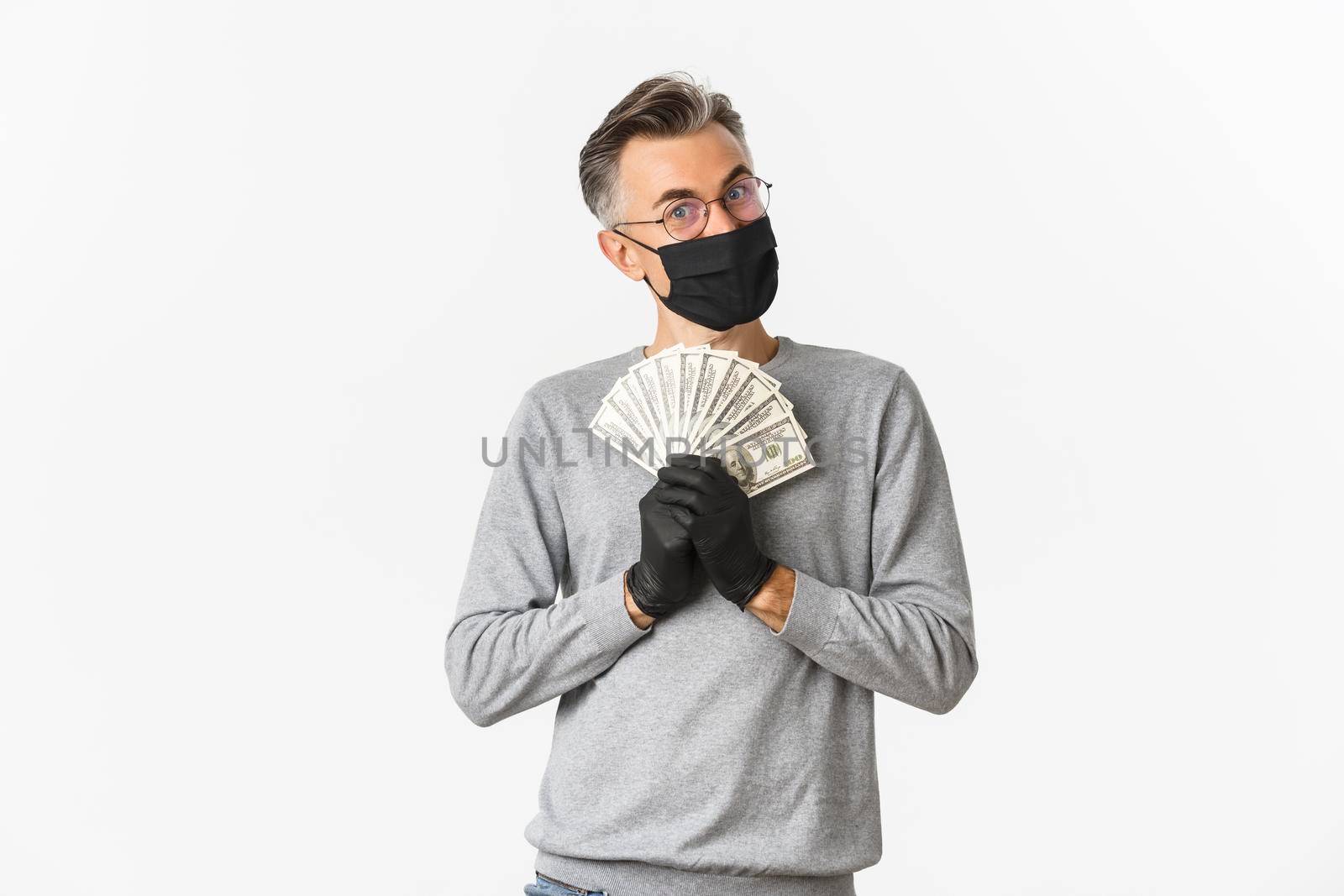 Concept of covid-19, social distancing and lifestyle. Portrait of cheerful rich man in medical mask, gloves and glasses, bragging with money and looking pleased, winning lottery.