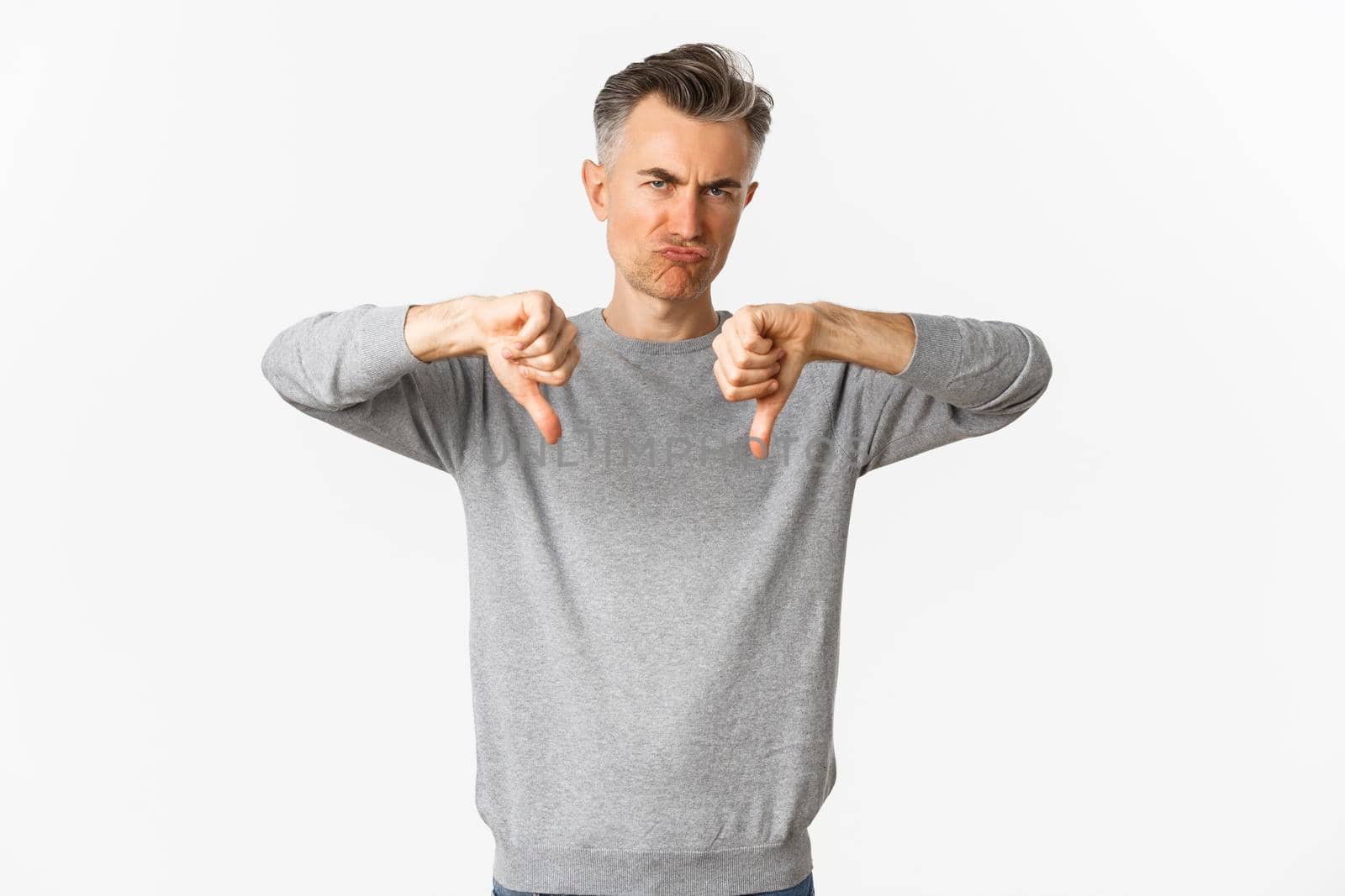 Skeptical and disappointed middle-aged man, grimacing unamused and showing thumbs-down, dislike something bad, standing over white background.