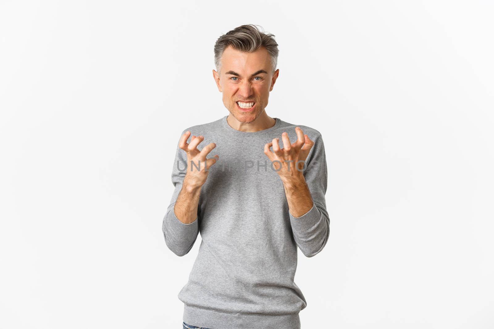 Image of angry middle-aged man cursing someone, looking mad and clenching fists with hatred, standing over white background.