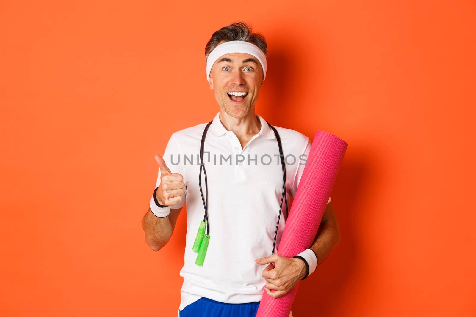 Concept of workout, gym and lifestyle. Cheerful middle-aged fitness guy, holding skipping rope and yoga mat, showing thumbs-up and smiling satisfied, standing over orange background.