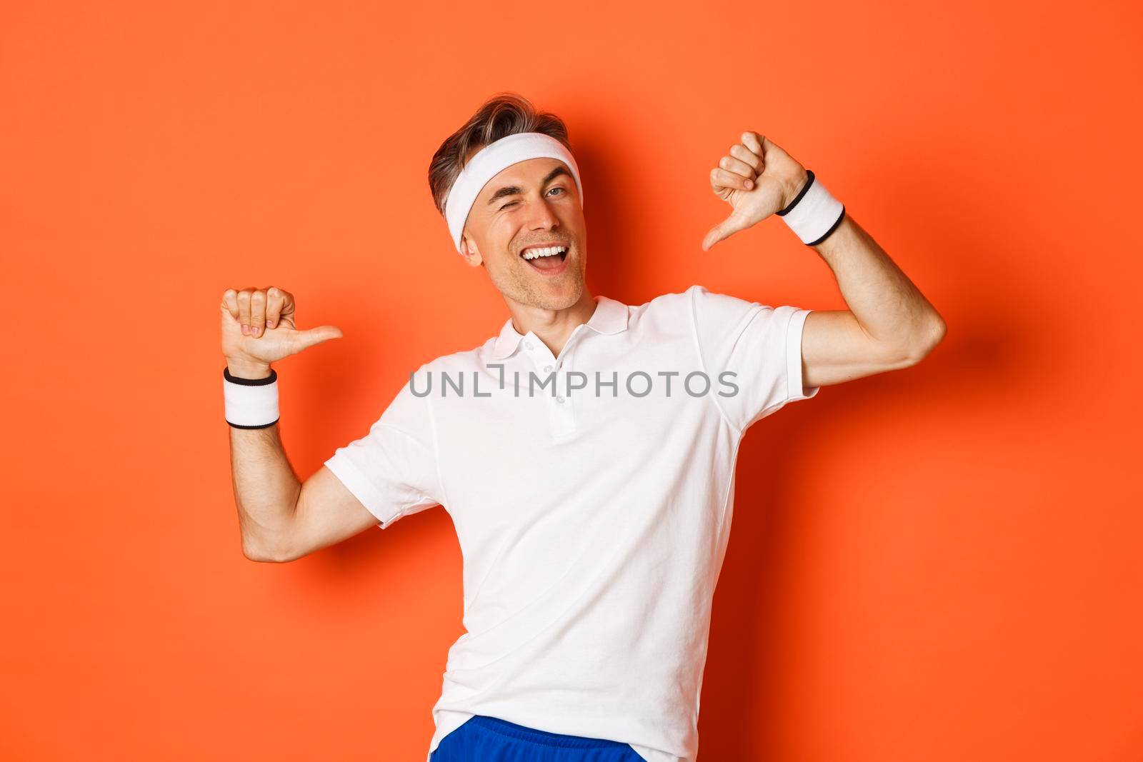 Concept of sport, fitness and lifestyle. Image of confident middle-aged man pointing at himself, bragging about achievement, wearing clothes for workout, standing over orange background.