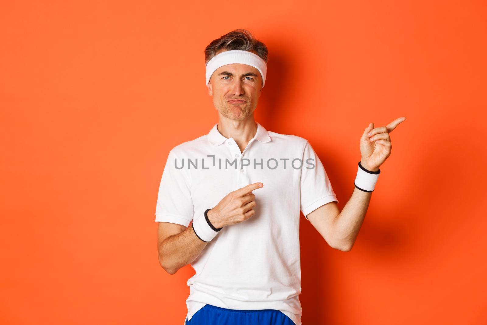 Concept of workout, sports and lifestyle. Portrait of skeptical senior sportsman in activewear, complaining about something bad, grimacing and pointing fingers right, orange background.