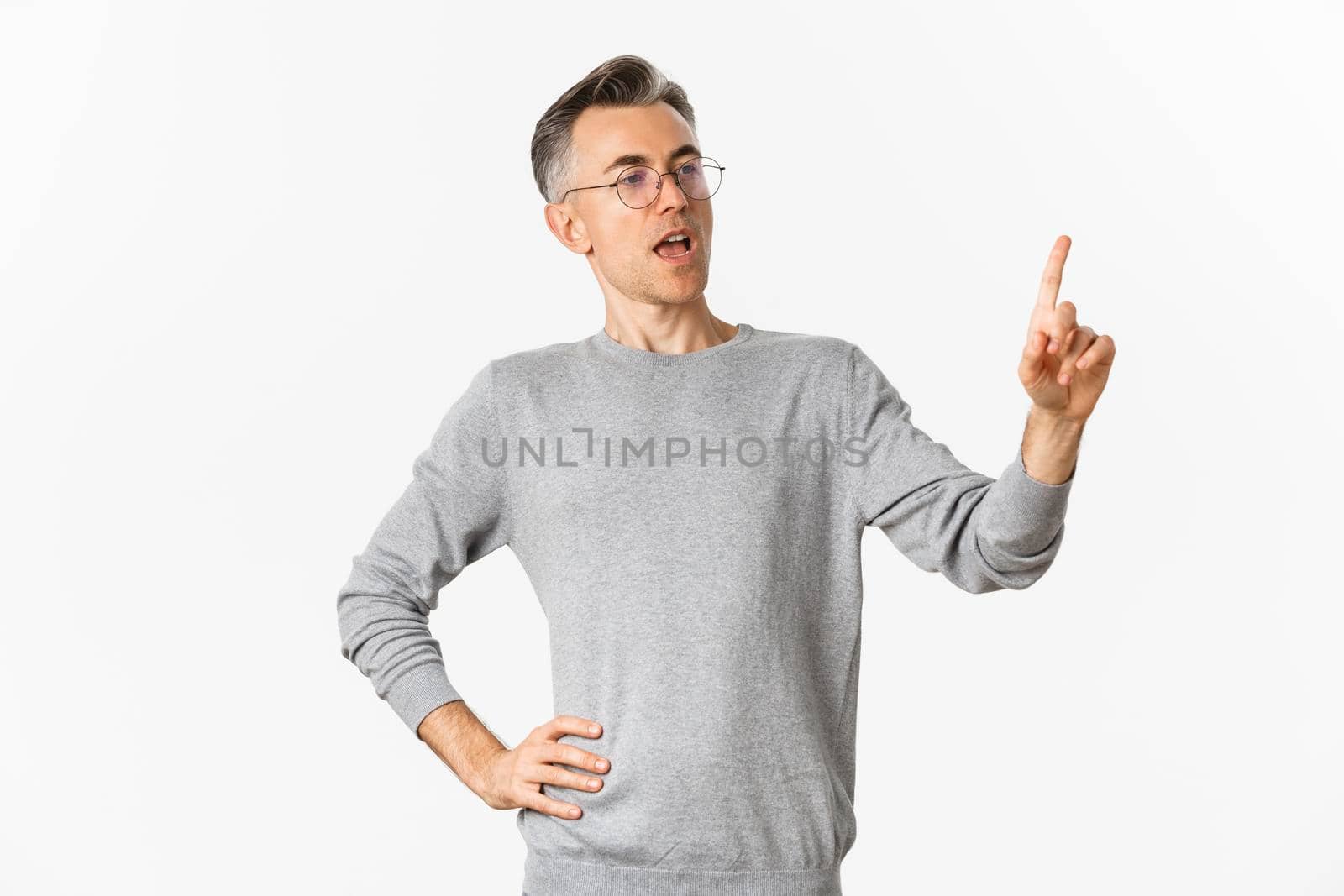 Image of confident middle-aged businessman in grey sweater and glasses, pointing finger and making speech, giving tasks to employees, standing over white background.