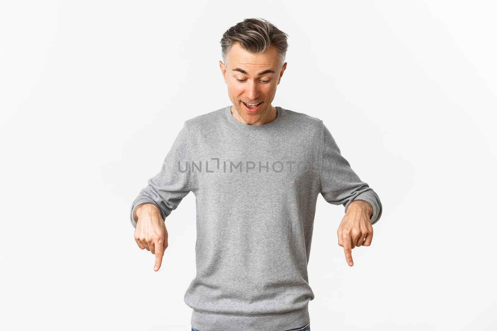 Image of excited middle-aged man in grey sweater, looking and pointing down with amazed face expression, standing over white background.