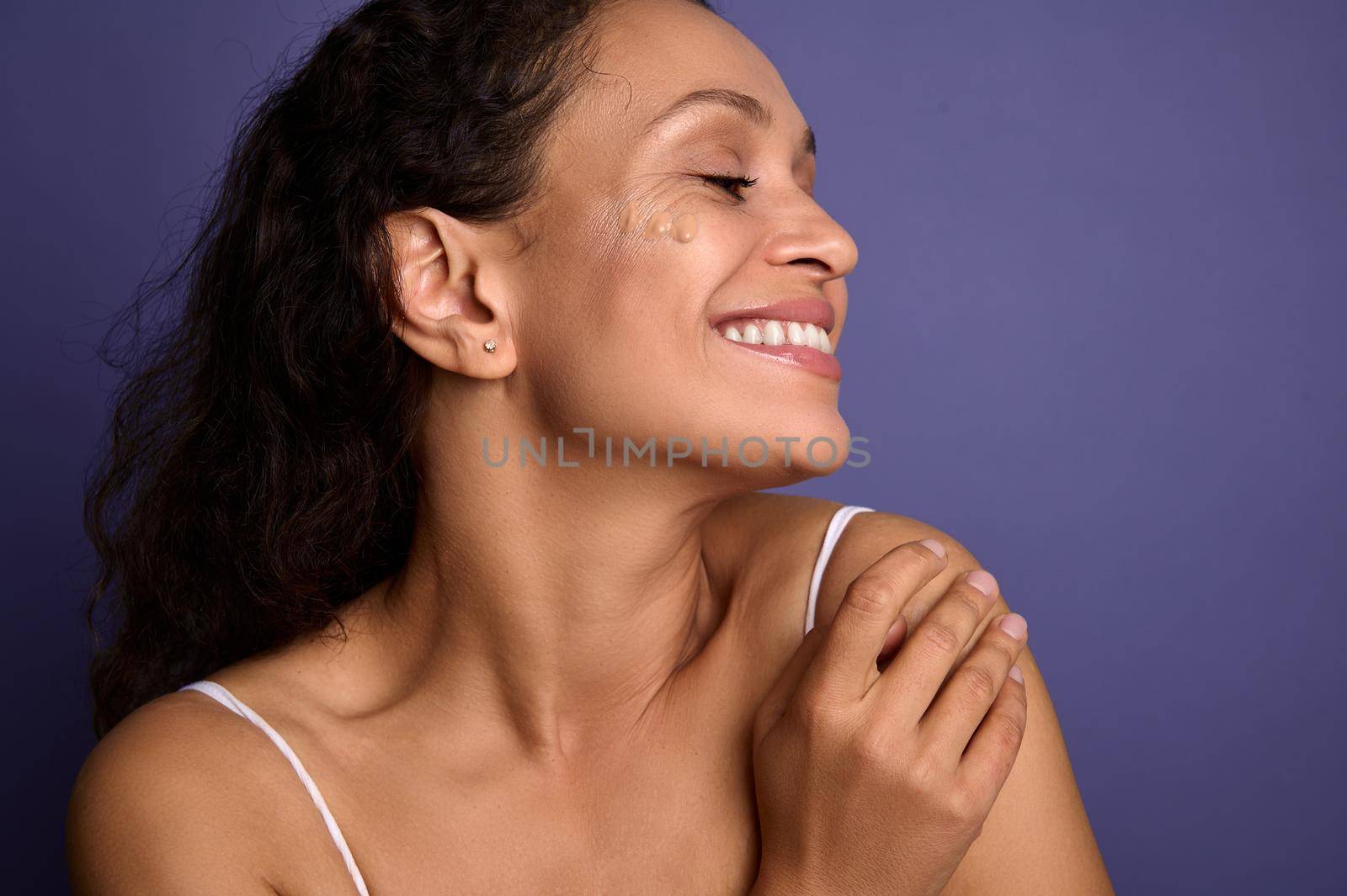 Beautiful dark-haired African woman with clean healthy glowing skin posing with spots of liquid tonal foundation on her face, smiles toothy smile looking aside over purple background with copy space. by artgf