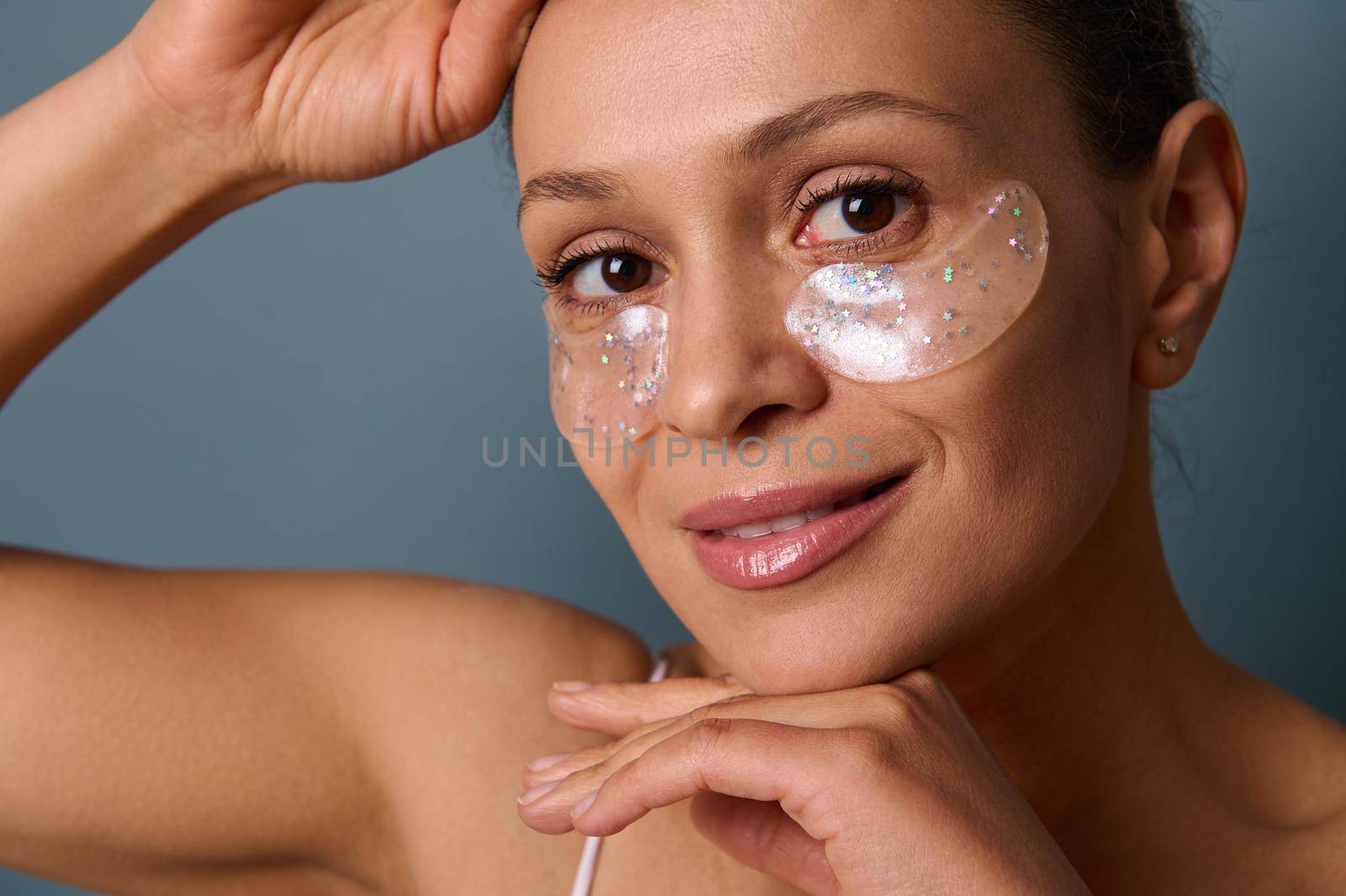 Close-up portrait of an attractive smiling woman applying gold medical hydrogel collagen eye patches to reduce bags under her eyes and puffiness, posing at camera against a gray background. Copy space by artgf