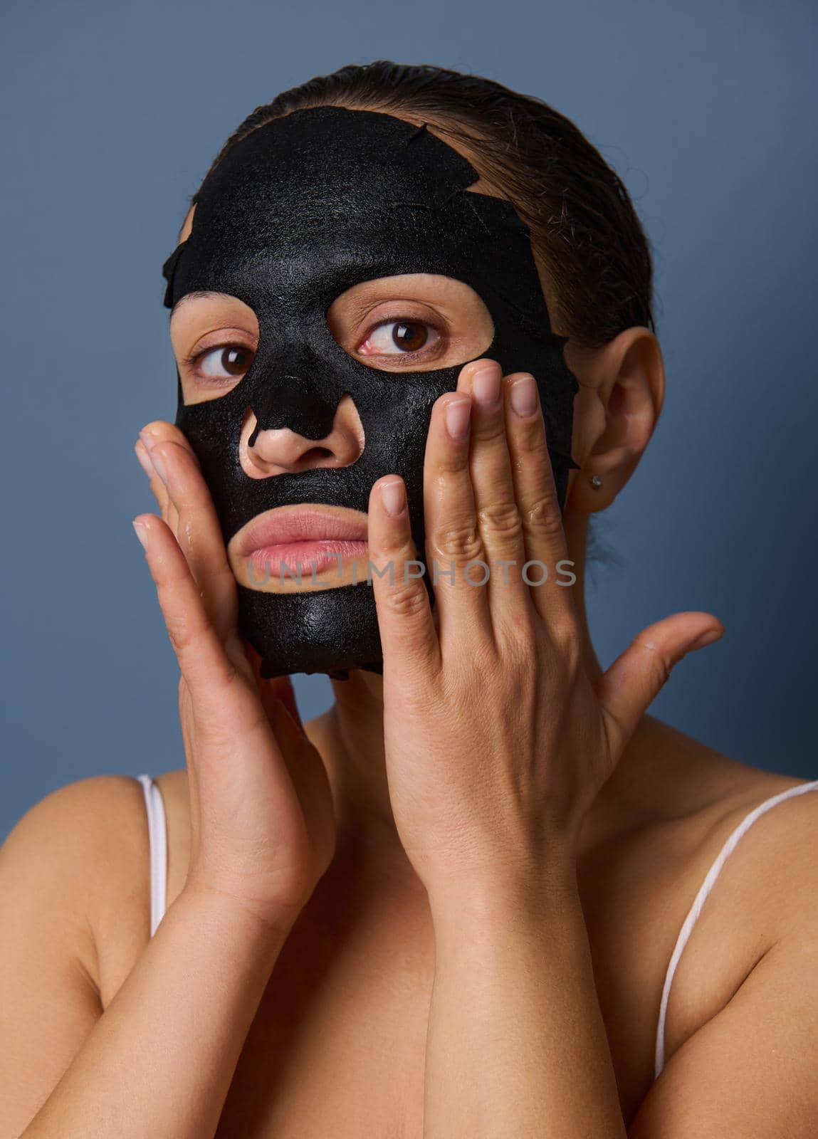 Close-up portrait of beautiful woman looking at the camera while putting on a nourishing smoothing moisturizing black face mask taking care of the beauty of her skin. Isolated on blue-gray background by artgf