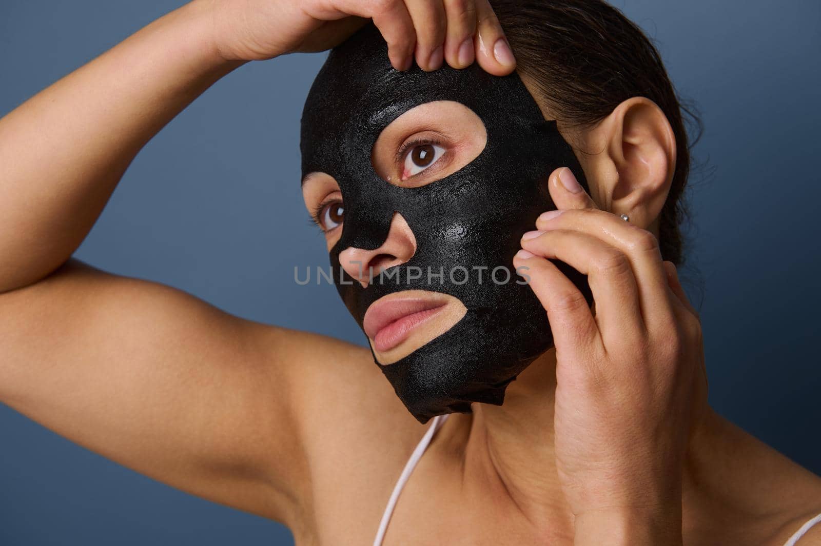 Close-up of beautiful woman putting on a hydrating smoothing nourishing cleansing black fabric mask on her face, isolated over gray background with copy space. Skin care, cosmetology, home spa concept