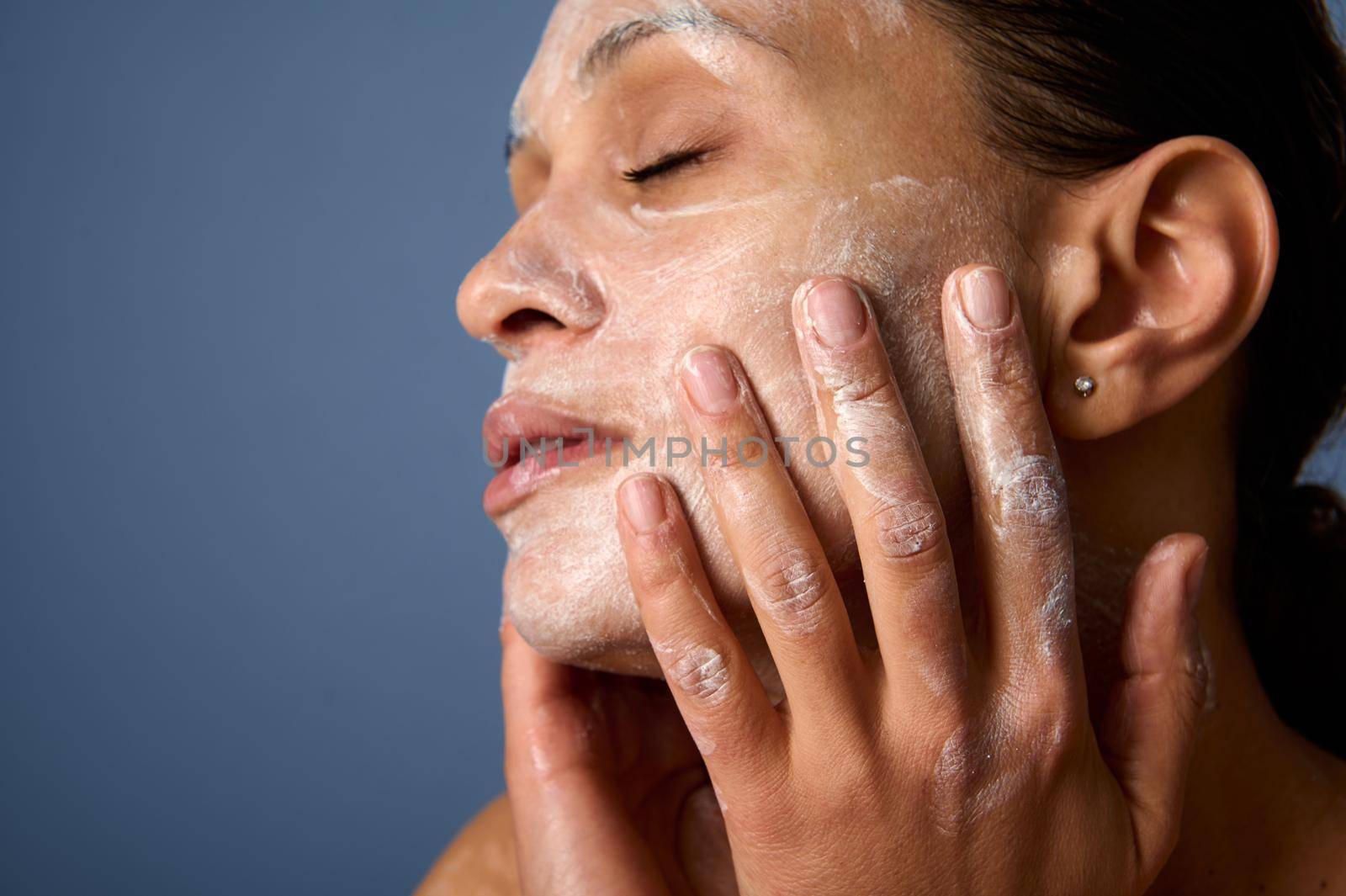Head shot of beautiful woman with perfectly healthy skin, holding hands on her face, performing smoothing massage, removing makeup and cleansing face with exfoliating scrub and facial foam cleanser by artgf