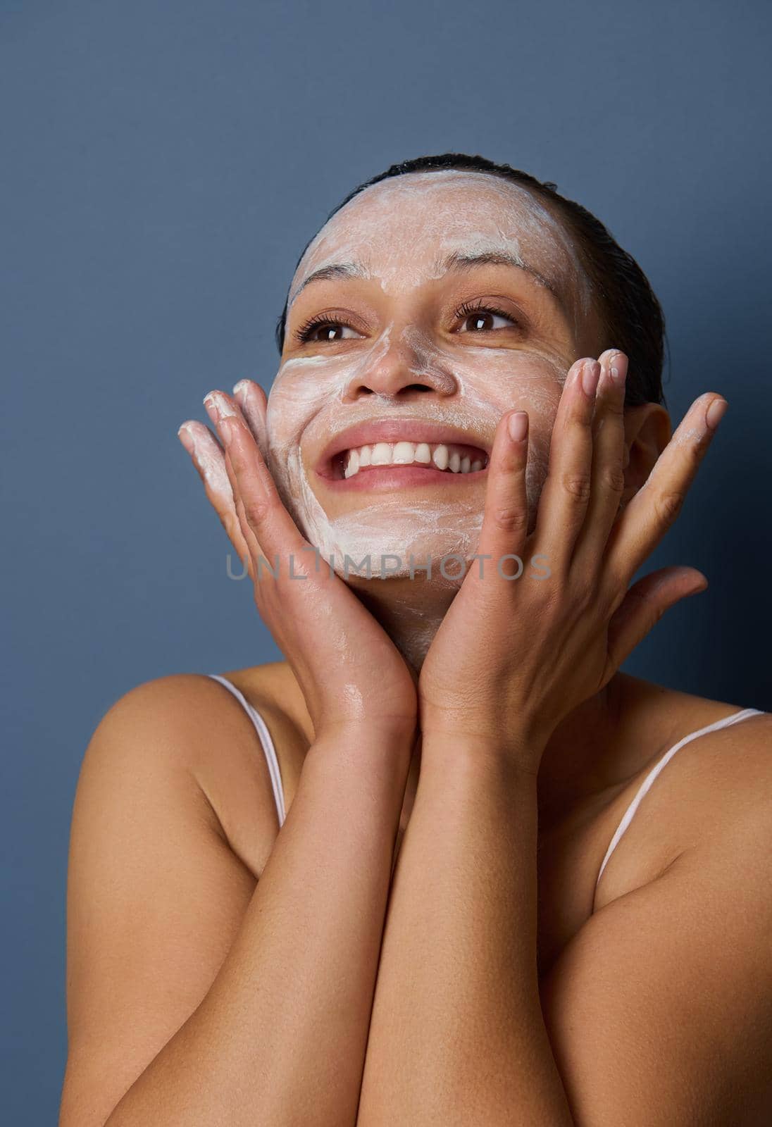 Beauty portrait of an attractive delightful wonderful woman with beautiful toothy smile and healthy skin removing make-up and cleaning her face with facial exfoliant scrub cleanser, on blue background by artgf