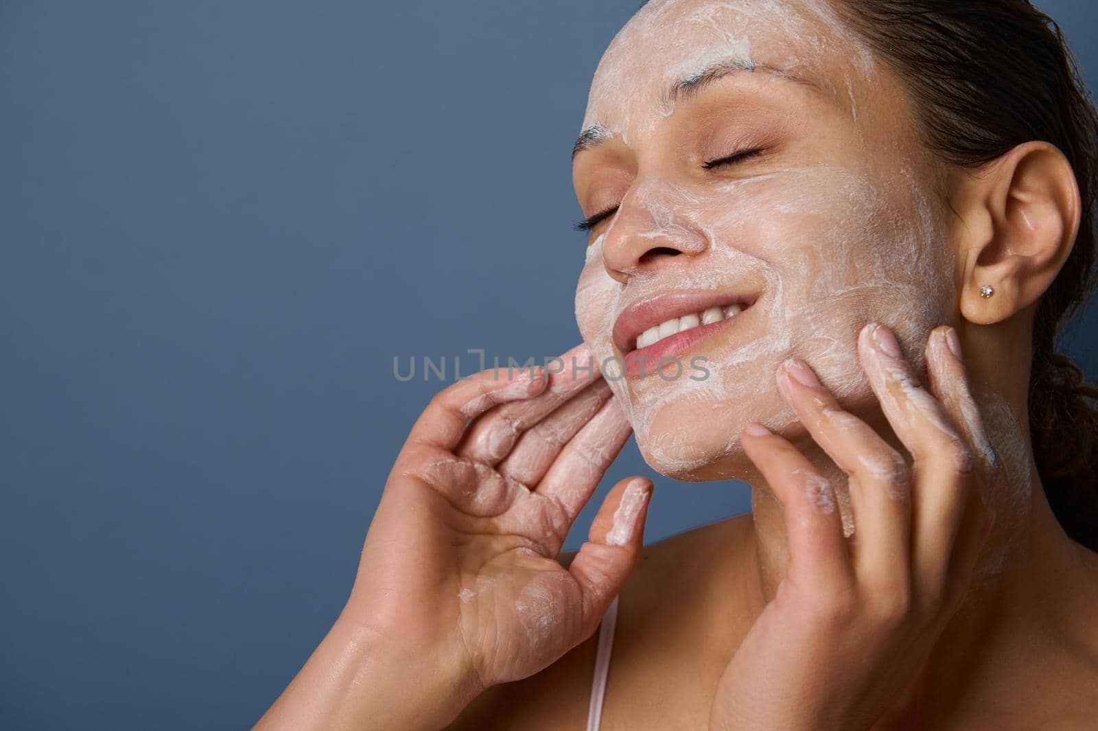 Beautiful delighted woman with perfectly healthy skin, holding hands on her face, performing smoothing massage, removing makeup and cleansing face with exfoliating scrub and facial foam cleanser. by artgf