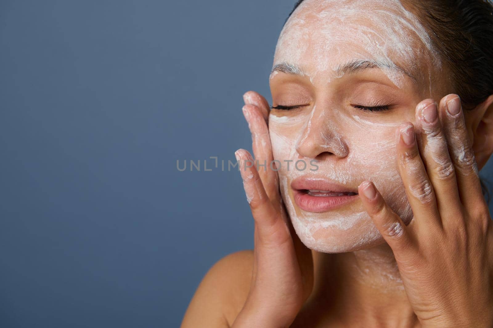 Close-up. Beautiful woman massaging her face while removing make-up using a foam cleansing cosmetic product, and refreshing her skin with an exfoliant beauty product, isolated over gray background