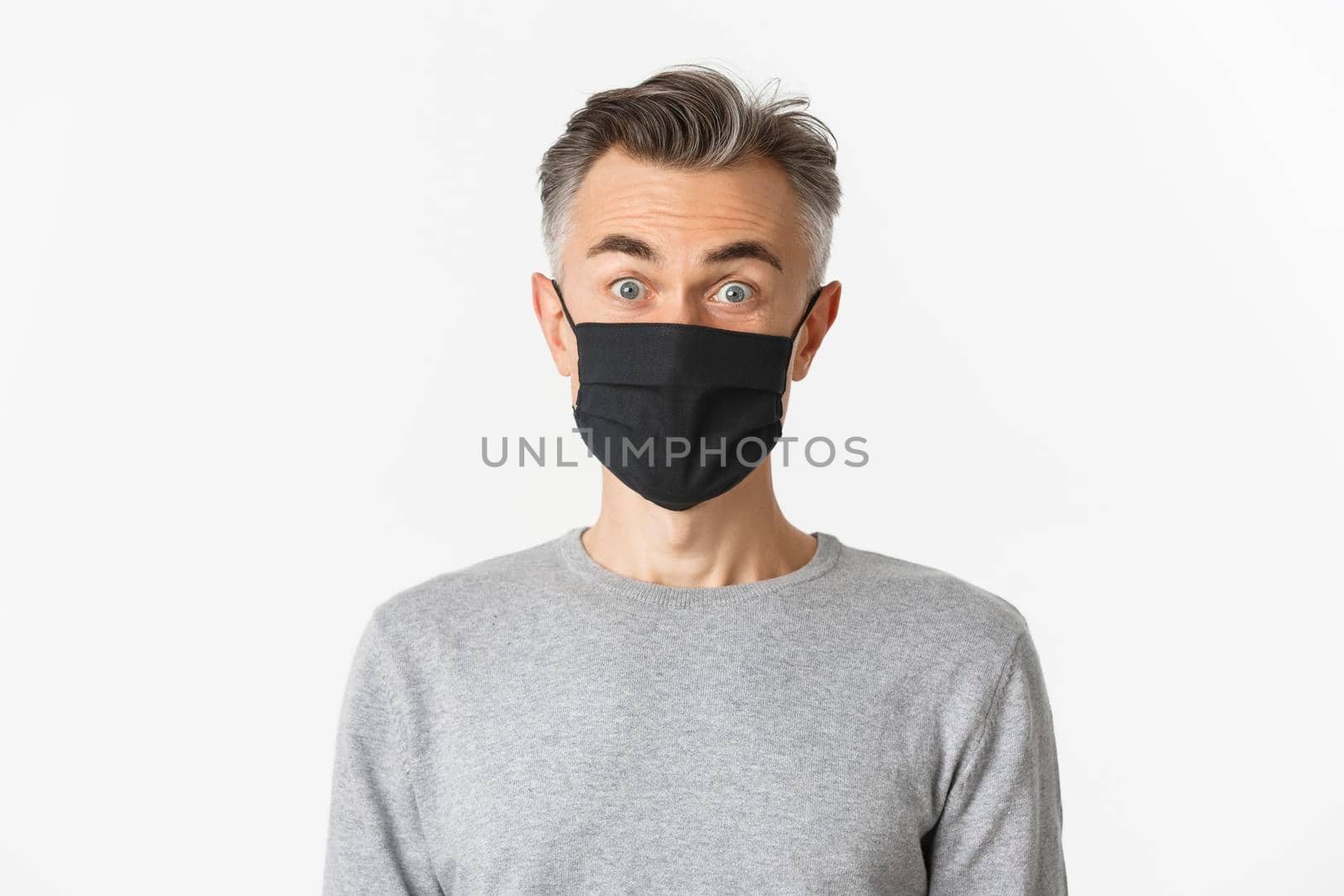 Concept of covid-19, social distancing and quarantine. Close-up of surprised middle-aged man in black medical mask, hear fantastic offer, look amazed at camera, standing over white background.