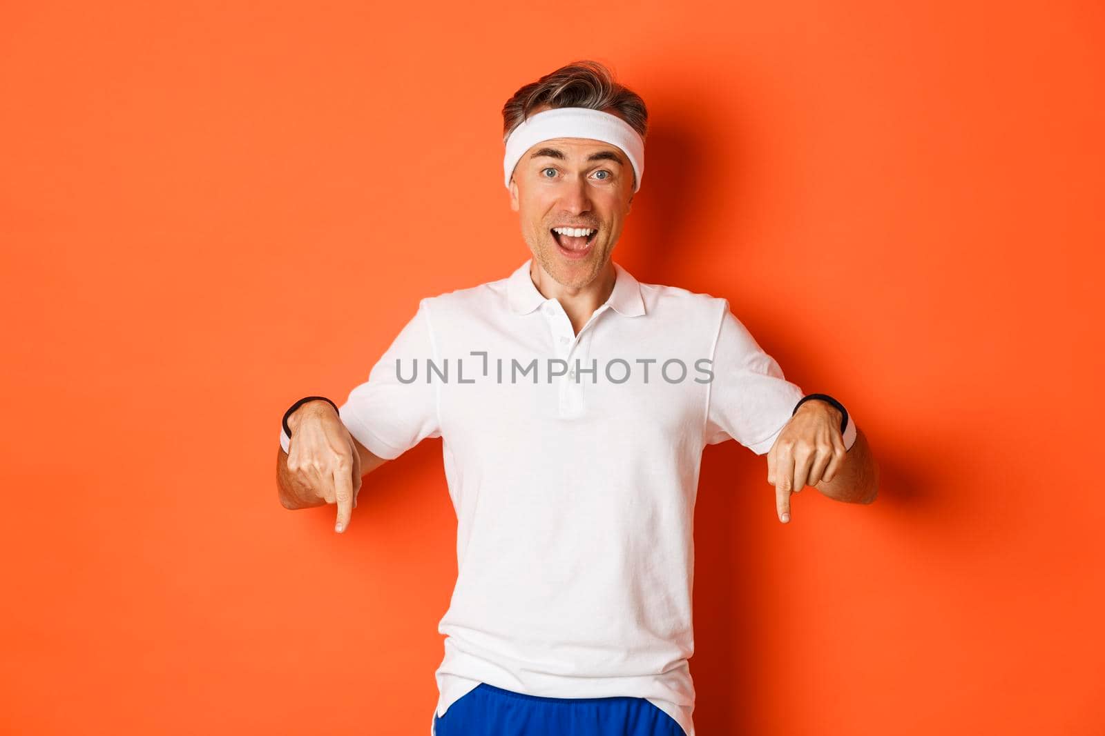 Concept of workout, sports and lifestyle. Portrait of happy middle-aged man in gym uniform, pointing fingers down and smiling amazed, showing promo offer, standing over orange background.