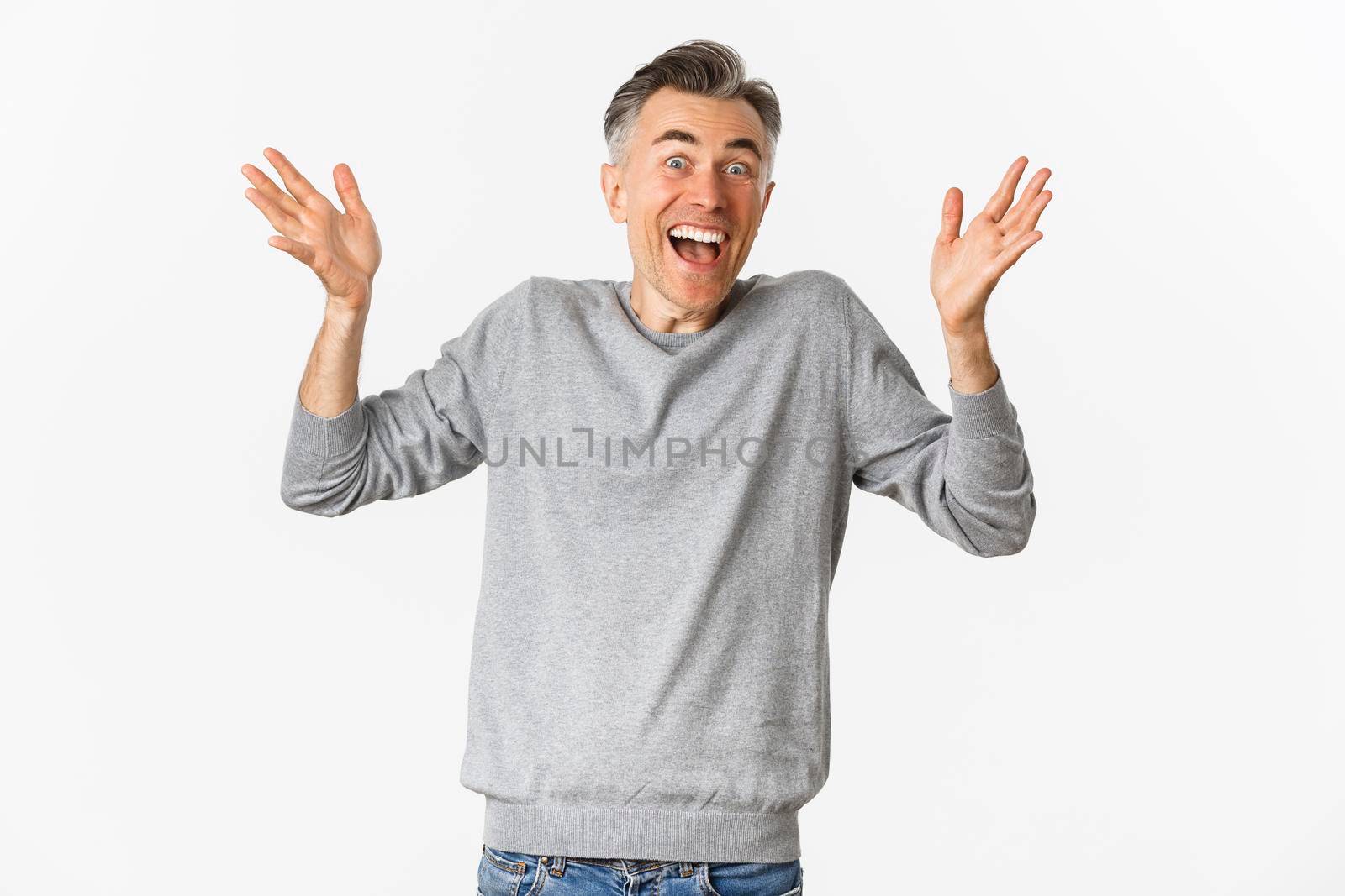 Image of excited and happy middle-aged man with gray short hairstyle, raising hands up and cheering, standing over white background.