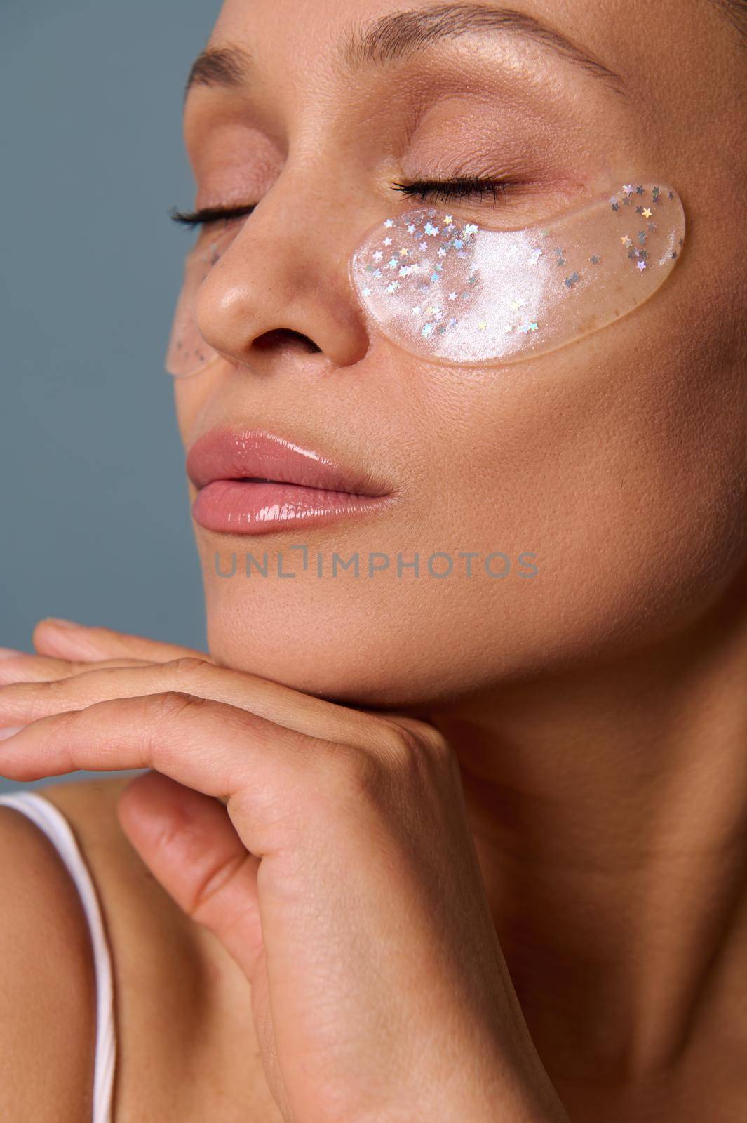 Close-up of the face of a beautiful woman with perfect glowing skin and natural makeup posing for camera with medical collagen patches under her eyes to reduce fatigue and puffiness. Skin care concept