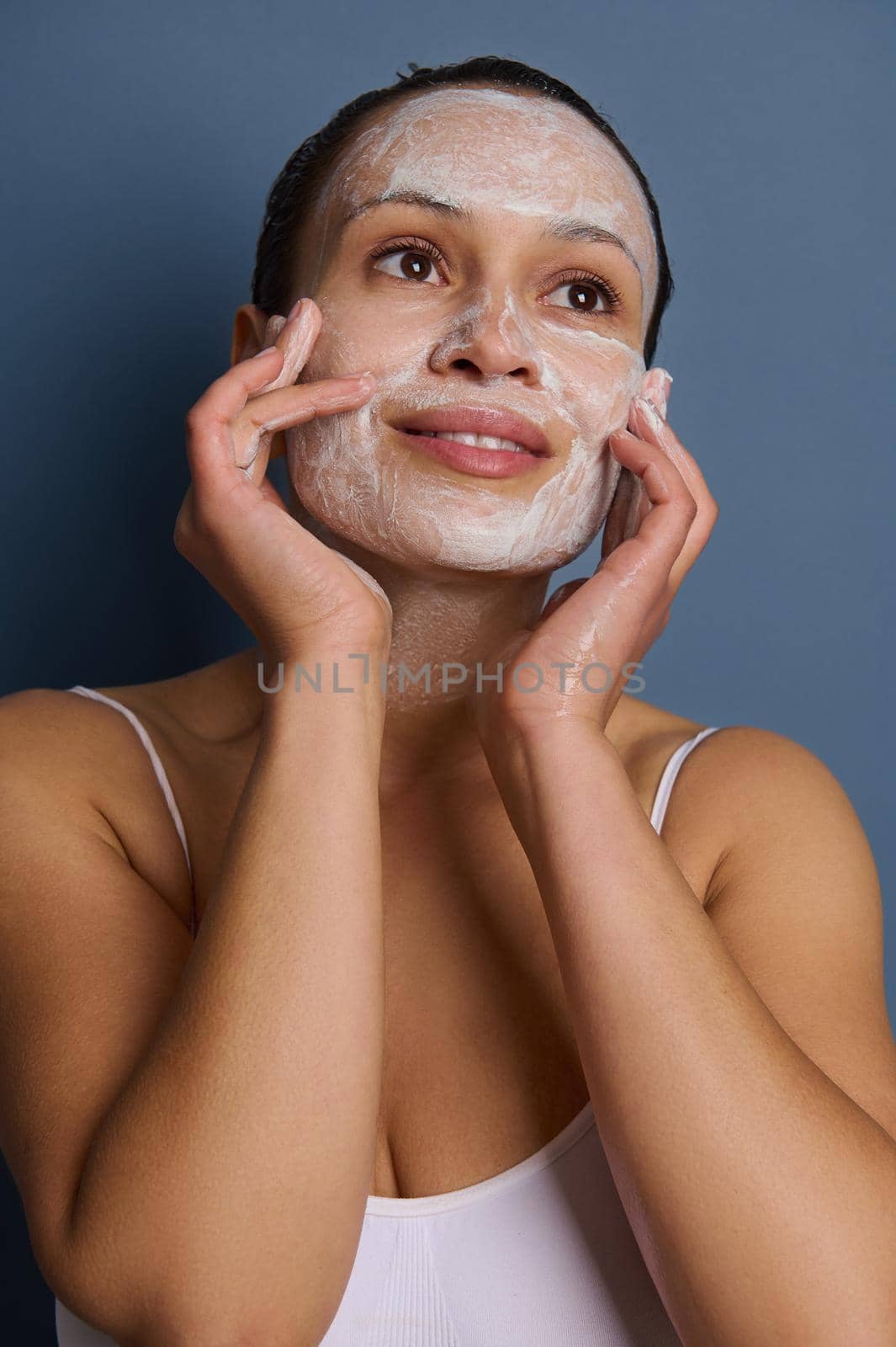 Beautiful woman applying foam cleansing cosmetic product on her face, doing massage movements, removing make-up and refreshing her skin using an exfoliant beauty product, isolated over gray background