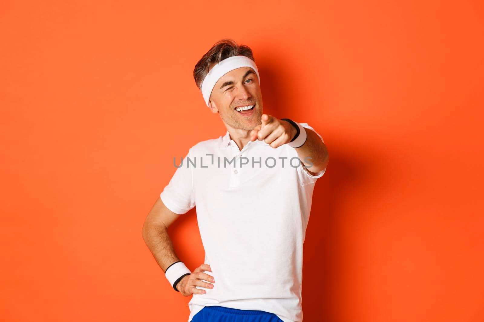 Concept of sport, fitness and lifestyle. Portrait of handsome, healthy middle-aged male athlete, pointing finger at camera and winking, inviting you to gym, standing over orange background.