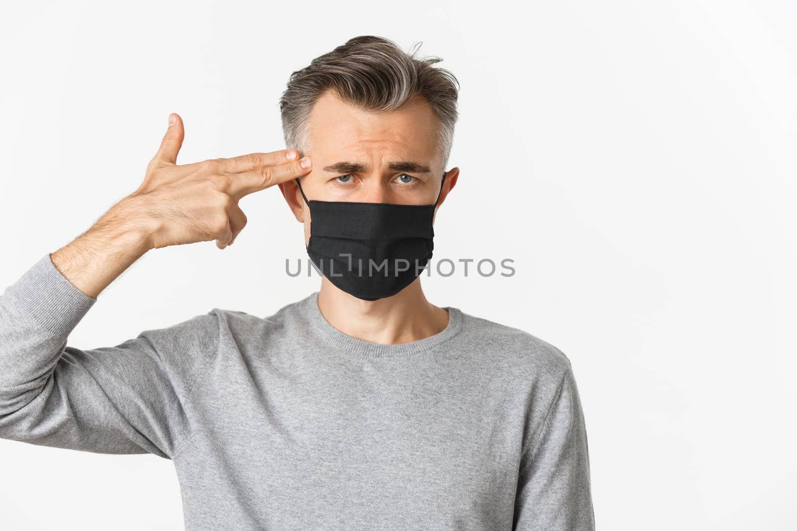 Concept of covid-19, social distancing and quarantine. Close-up of sad middle-aged man in black medical mask, pointing finger gun at head and looking distressed, standing over white background.