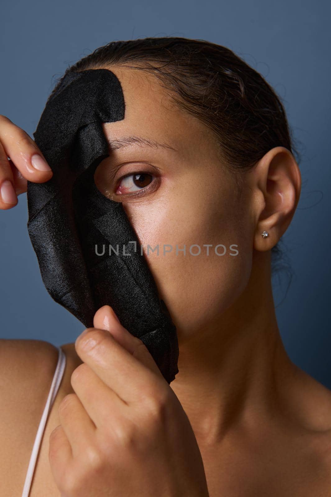 Close-up portrait of an attractive beautiful woman with natural make-up and fresh glowing healthy skin taking off fabric cosmetic black peeling mask from her face skin. Cosmetology, spa beauty therapy