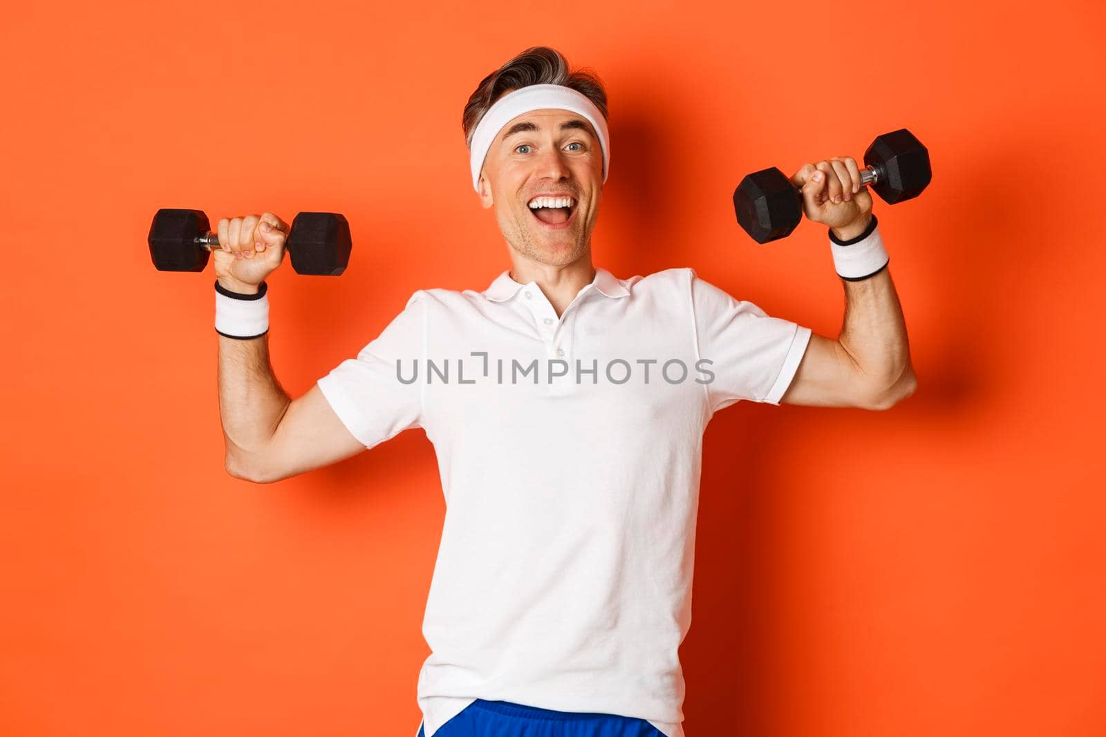 Concept of workout, gym and lifestyle. Image of healthy and strong middle-aged male athlete, doing sport exercises with dumbbells and smiling, standing over orange background.