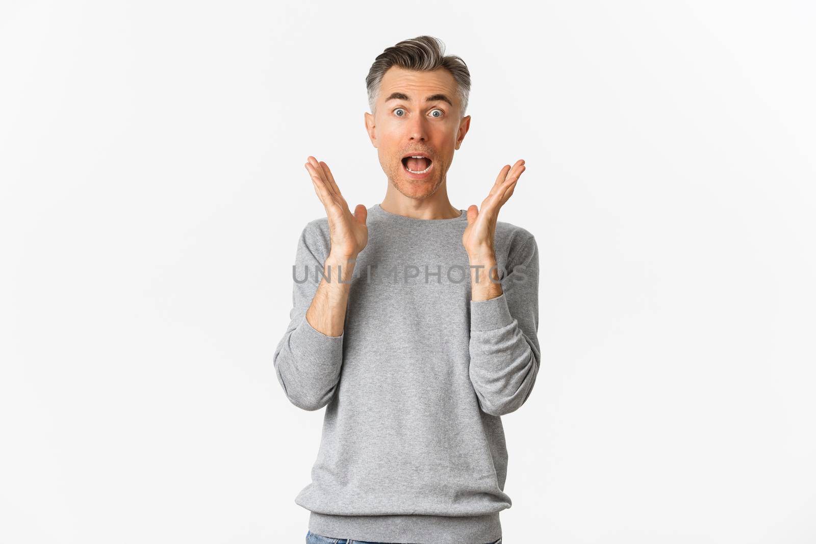 Surprised adult man with gray hair, saying wow and looking amazed, standing near white copy space in basic sweater.