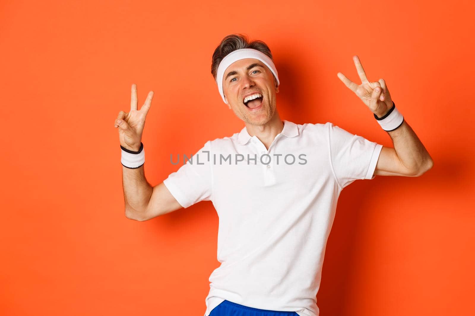 Image of happy and healthy middle-aged man in sport clothing, showing peace signs and smiling, workout over orange background.