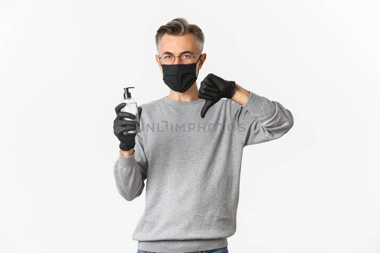 Concept of coronavirus, lifestyle and quarantine. Portrait of middle-aged man in medical mask and gloves do not recommend antiseptic, showing hand sanitizer and thumbs-down, white background.