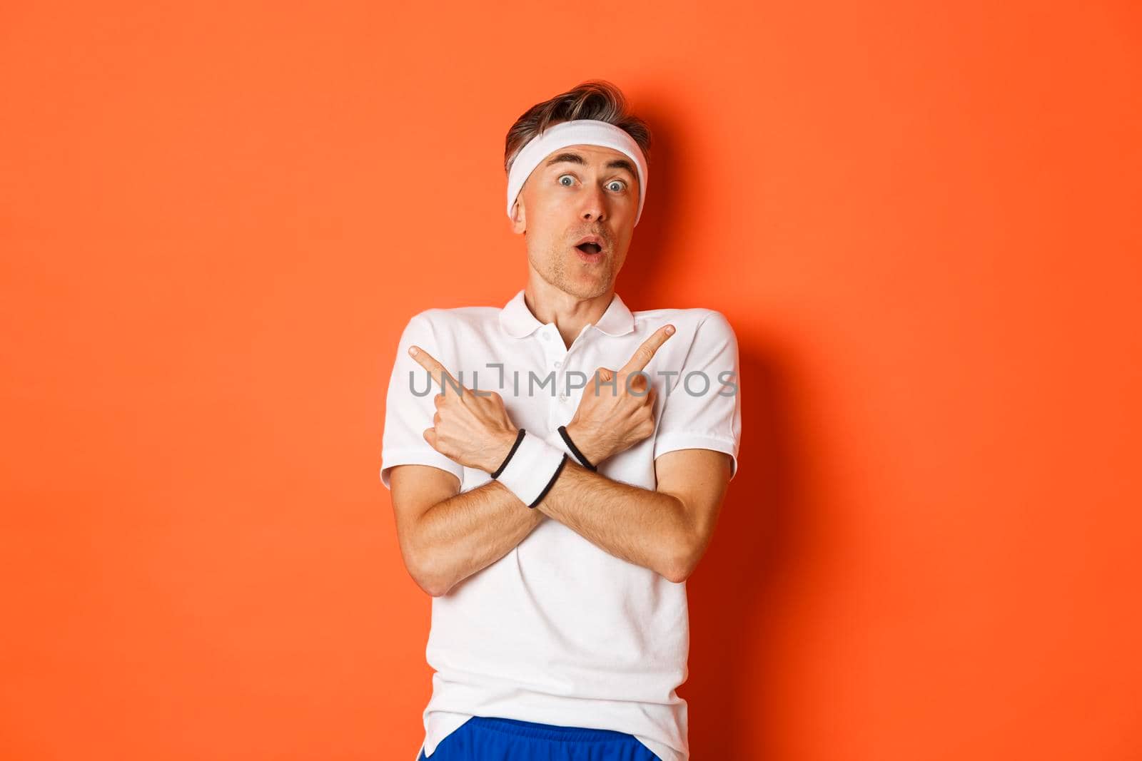 Concept of sport, fitness and lifestyle. Portrait of startled middle-aged male athlete, pointing fingers sideways and looking amazed, standing over orange background.