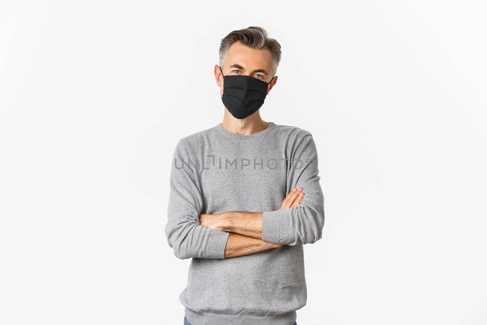 Covid-19, pandemic and social distancing concept. Middle-aged man with grey short hairstyle, wearing black medical mask, standing with hands crossed on chest against white background.