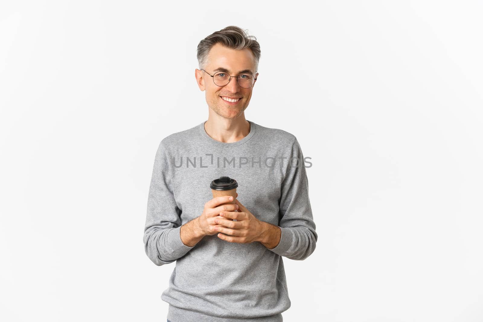 Image of handsome middle-aged man with grey short hair, wearing glasses and grey sweater, drinking coffee and smiling pleased, standing over white background.