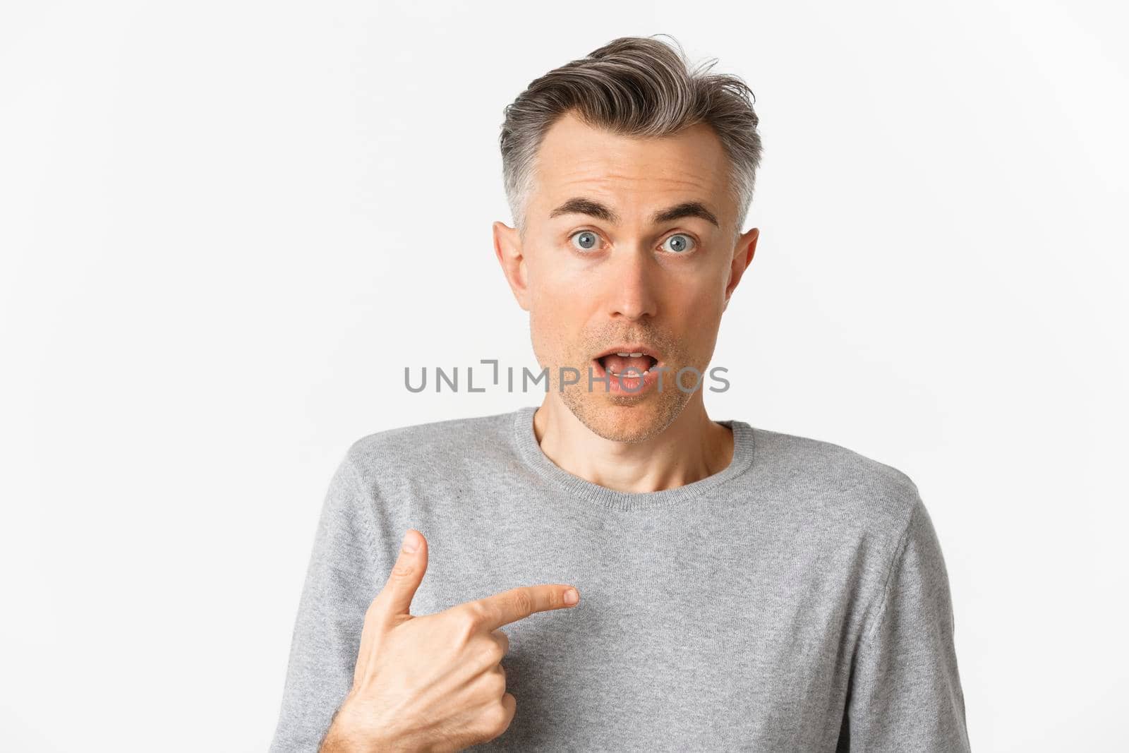 Close-up of confused middle-aged man, pointing at himself and gasping, standing over white background.