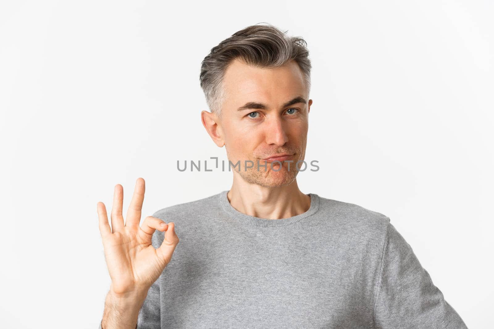 Close-up of satisfied middle-aged man in gray sweater, showing okay sign in approval, praising something good, standing over white background.