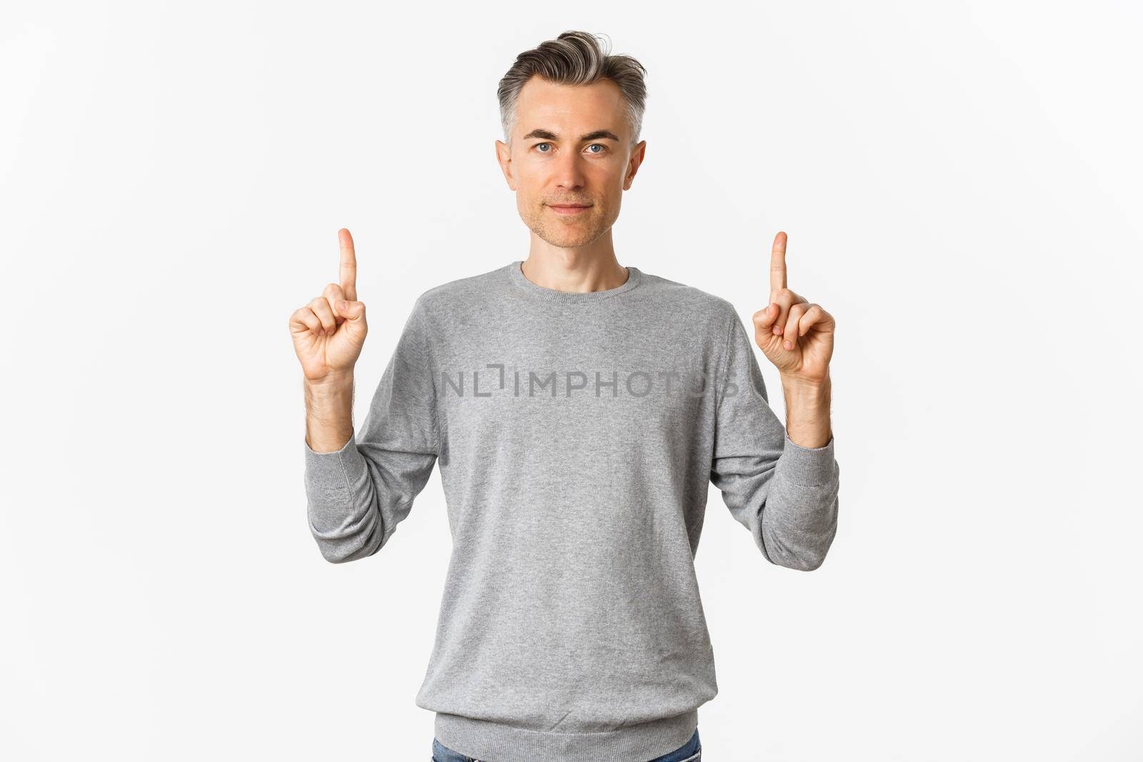 Portrait of handsome middle-aged man in grey sweater, showing promo, pointing fingers up and looking serious, standing over white background.