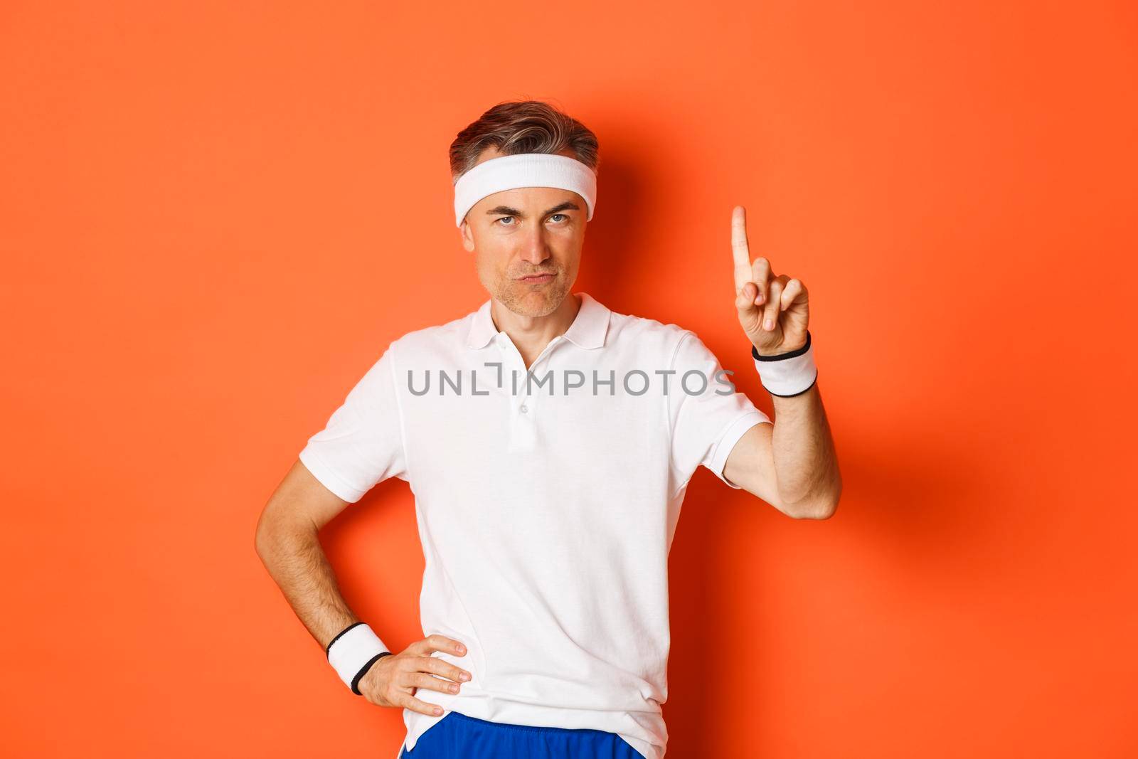 Portrait of serious middle-aged male athlete, wearing sportswear, pointing finger up and showing logo about sports and gym, standing over orange background.