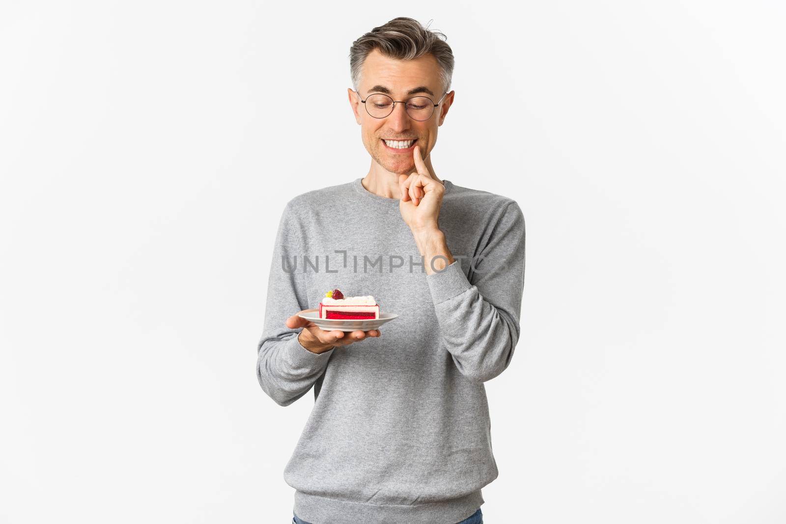 Portrait of handsome middle-aged man in glasses and gray sweater, looking tempted at delicious cake, wanting to eat dessert, standing over white background.