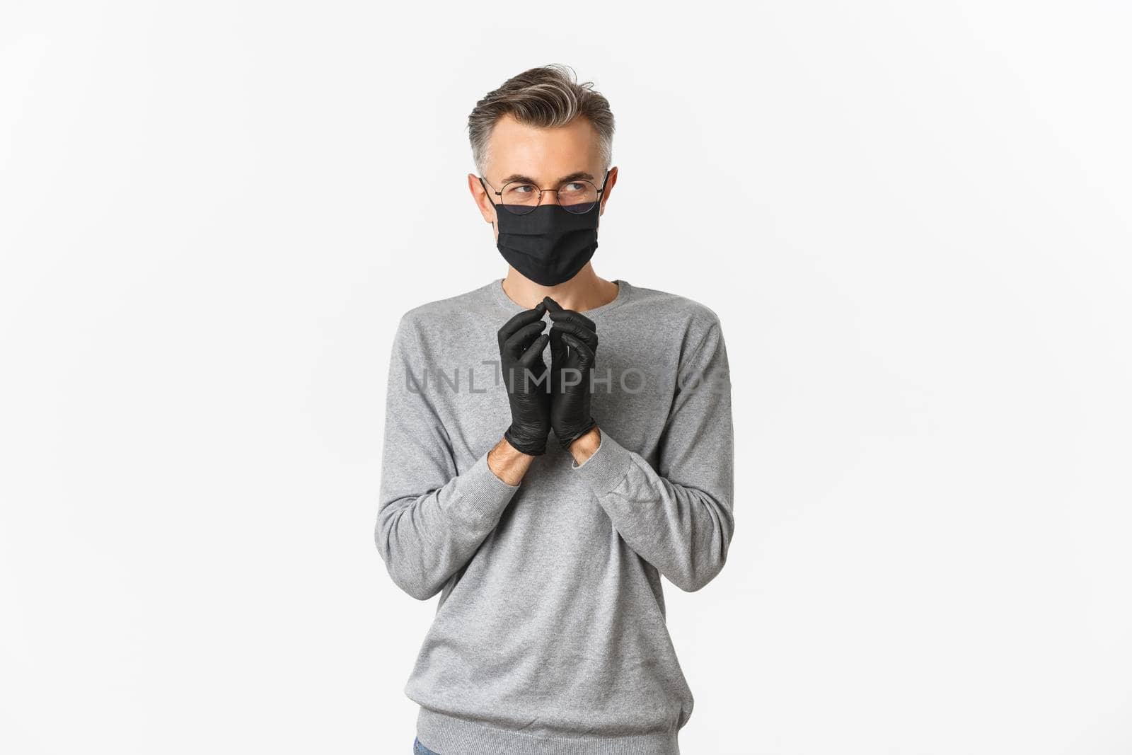 Concept of covid-19, social distancing and lifestyle. Image of cunning middle-aged man in black medical mask and gloves, making plan, scheming something bad, standing over white background.